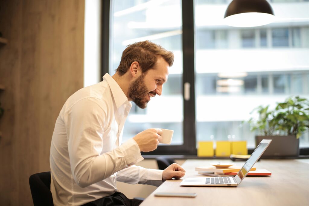 pexels photo 925786 925786 Man in office with coffee, smiling while working at laptop, captures the essence of remote work.